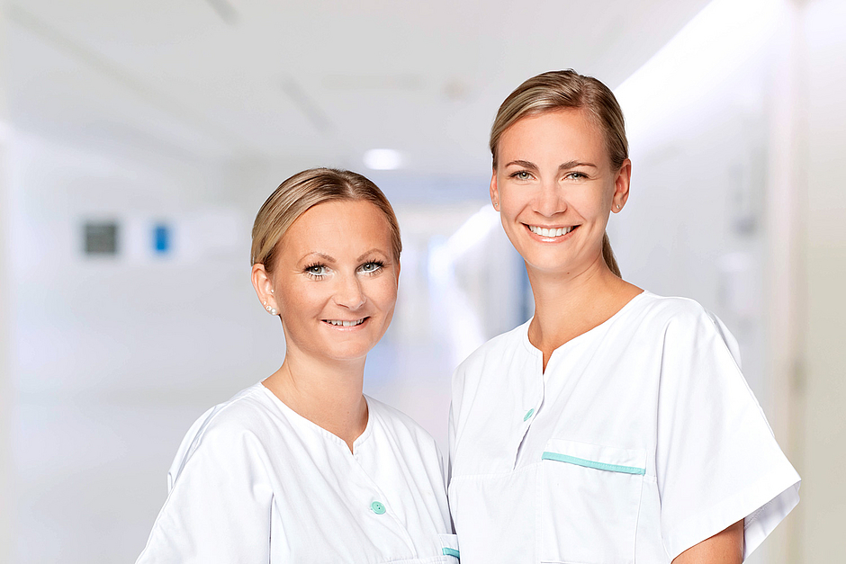 Zwei Kolleginnen auf dem Klinikflur schauen gemeinsam in die Kamera